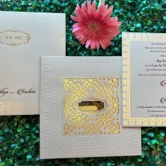 An elegant white and gold invitation featuring a detailed laser-cut 'jaali' window. The intricate pattern creates a beautiful play of shadow and light.