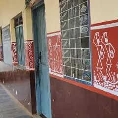 A view down the school corridor, showing how the Warli murals are integrated between the doors and windows. The paintings depict scenes like women drawing water from a well, adding narrative details to the building's exterior.