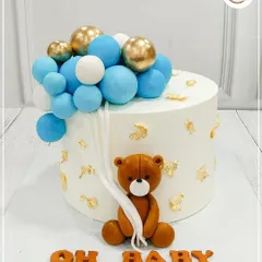 A classic "Oh Baby" cake featuring a cute teddy bear holding a string of blue and gold balloons. The gold leaf accents add a touch of elegance.