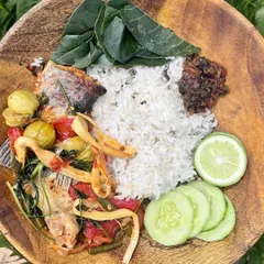 A simple, wholesome farm-style meal. This wooden bowl holds a comforting mix of rice, fish curry with bitter eggplant and foraged greens, and a side of fresh cucumber and lemon.