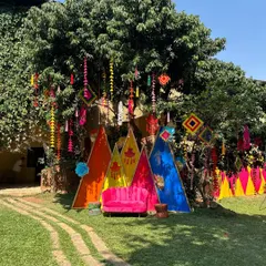A wide view of the Mehandi setup, featuring a colorful triangular backdrop and traditional hangings from a large tree. The entire space was designed to feel playful and full of celebratory energy.