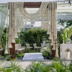 An open-air mandap designed for a serene and beautiful ceremony. The structure is made of polished teak pillars, draped with cascading strings of fresh tuberose for a fragrant and visually stunning effect.