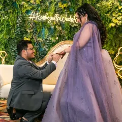 The groom places the ring on his bride's finger during their ring ceremony, set against a lush green wall with a custom neon sign.