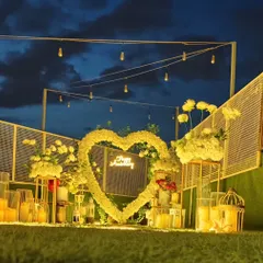 An elegant rooftop anniversary celebration at dusk. A heart-shaped arch of white flowers frames a "Happy Anniversary" neon sign, with string lights overhead and candles all around creating a sophisticated and romantic mood.