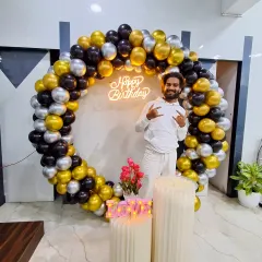 A customer posing with the black, gold, and silver balloon ring. This shows the scale of the backdrop, which features a 'Happy Birthday' neon sign.