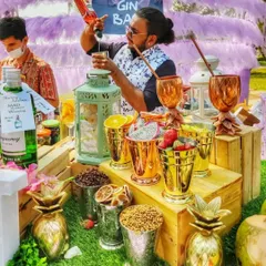 An action shot of a bartender crafting a custom gin cocktail. He pours a vibrant red mixer into a glass, surrounded by an array of fresh garnishes like oranges, dragon fruit, and herbs in metallic cups.
