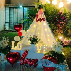 An outdoor cabana tent setup for a romantic 'LOVE' themed celebration. The decor includes light-up letters, heart-shaped balloons, and floral arrangements.