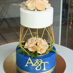 A bold and modern wedding cake design. The royal blue bottom tier with gold initials supports a geometric gold separator, creating a 'floating' effect for the clean white top tier, all adorned with fresh flowers.