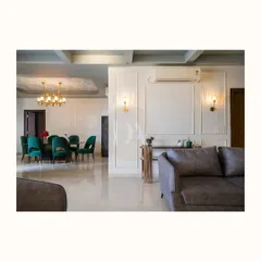 Here is a glimpse into an open-plan living and dining area. The design connects the two zones seamlessly, using classic white wall paneling and elegant sconces for a cohesive look. The emerald green dining chairs add a rich pop of color against the neutral backdrop.