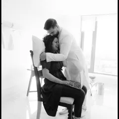 A sweet black and white photo of the groom hugging the bride from behind while she works on her laptop, a real-life moment of love amidst wedding planning.