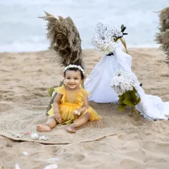 A sweet photo of a baby girl sitting on a jute mat on the beach for her first birthday, with the waves gently rolling in behind her.