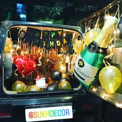 For a fun and unique surprise, I offer car boot decorations. This setup features a 'Happy Birthday' banner, a shimmering backdrop, festive gold and black balloons, and a large champagne bottle foil balloon to get the celebration started anywhere.