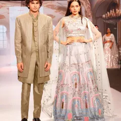 Showcasing a modern, coordinated look for a stylish couple. The groom wears a unique layered Indo-western outfit that perfectly complements the bride's pastel lehenga with a cape-style dupatta.