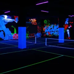 A wide-angle view of the entire pickleball court glowing under UV blacklights. The neon lines of the court and the glowing murals work together to create a completely immersive and futuristic playing environment.