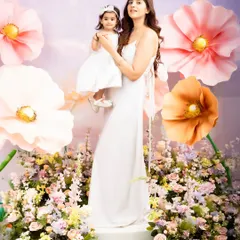 A full-length portrait of a mother and daughter in a beautiful, flower-filled studio setup. Their matching white dresses and the whimsical background create a stunning, high-fashion-inspired image.