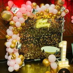 A chic and modern first birthday setup. The decoration is built around a gold shimmer wall, a "Happy Birthday" neon sign, a light-up number "1", and a pink and gold balloon arch.