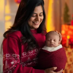 A smiling mother holds her swaddled newborn close in a warm, festive setting with Christmas lights.