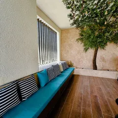 This cozy balcony nook features a long built-in bench with bright turquoise cushions. An artificial olive tree planted in a bed of pebbles adds a touch of Mediterranean charm and natural shade.