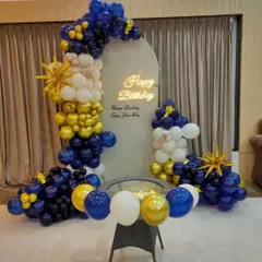 A stylish birthday setup with a white arched backdrop. The decoration features a balloon garland in blue, yellow, and white, with starburst foil balloons and a neon sign.