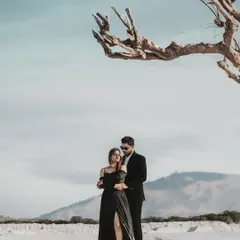 A dramatic and modern pre-wedding portrait set against a stark, beautiful landscape. The composition uses a bare tree to frame the couple, creating a powerful, high-fashion image that feels both artistic and romantic.