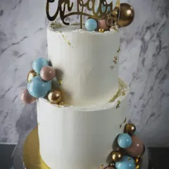 A two-tiered baby shower cake that’s both modern and elegant. The clean white buttercream is decorated with pink, blue, and gold chocolate spheres that look like balloons, perfect for a stylish celebration.