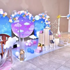 A dreamy 'Star and Moon' themed party setup. The decor features a night-sky backdrop with cloud and star cutouts, a crescent moon prop, and a beautiful balloon arrangement in shades of blue, pink, and gold.
