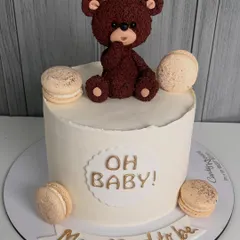 A simple and elegant "Oh Baby!" cake for a baby shower. This design features a textured teddy bear topper and is accented with delicious macarons, perfect for celebrating the mom and dad-to-be.