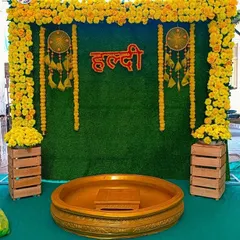 A simple yet beautiful Haldi decoration with a green grass backdrop, a frame of marigold flowers, and a neon Hindi "Haldi" sign. The wooden crates and dreamcatchers add a rustic, bohemian touch to the setup.