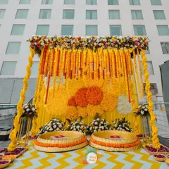 A classic Haldi ceremony setup, featuring a backdrop of marigold garlands, traditional low seating (urlis), and a bright yellow and white chevron floor.