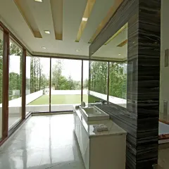 The master bathroom at Sachdeva Farmhouse is a light-filled space with floor-to-ceiling glass offering views of the pool and gym terrace. A sleek stone feature wall screens the sunken tub, creating a refined, spa-like feel.