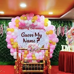 A fun and interactive naming ceremony decoration. The "Guess My Name?" backdrop, surrounded by a pink and gold balloon circle and a lush green wall, adds a playful element to the traditional event.