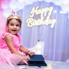 The birthday girl, dressed as a little fairy, sits happily at a table in front of a 'Happy Birthday' sign and balloon arch.