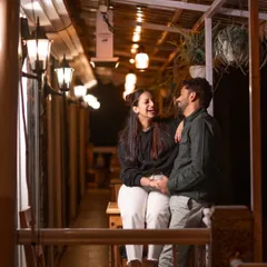 A relaxed evening moment on a beautifully lit verandah. The warm string lights create a cozy and inviting atmosphere for Ishu and Arihant's shared laughter, showing that romantic shots can also be casual and comfortable.