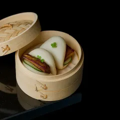 Our BBQ Pork Bao features delectable steamed buns filled with roasted pork, crisp cucumber slices, and our house BBQ sauce, served in a traditional bamboo steamer.