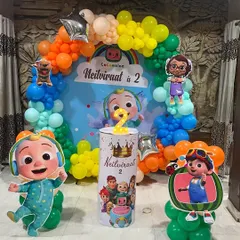 A vibrant Cocomelon theme decoration for a second birthday. The setup features a colorful rainbow balloon arch, a personalized backdrop, and standee cutouts of popular Cocomelon characters.