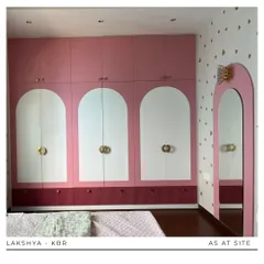The centerpiece of this girl's room is the custom wardrobe, featuring a playful design with pink and white arched panels and unique gold hardware. The heart-patterned wallpaper adds a touch of whimsy.