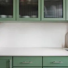 The kitchen in our Collectors House features custom cabinetry in a soothing sage green. Fluted glass on the upper cabinets adds a layer of texture and visual interest, partially obscuring the contents for a clean look.