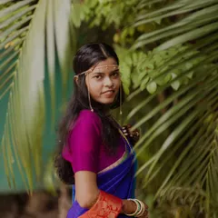 A beautiful portrait of a Maharashtrian bride in a vibrant blue and purple saree. Her simple yet elegant headpiece and joyful expression are perfectly framed by lush green leaves.