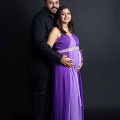 A classic maternity couple portrait. The parents-to-be stand together, smiling at the camera, showcasing their coordinated formal outfits against a timeless dark studio background.