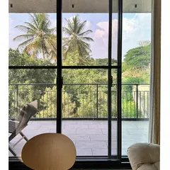 A view from inside the Jayanagar Residence, looking out through floor-to-ceiling glass doors to a balcony and the lush treetops beyond. This design choice effectively erases the boundary between the living space and the landscape.