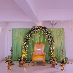 An elegant Valaikappu or Seemantham (baby shower) setup. The decor features a lush floral arch in white and yellow against a soft green drape, with traditional brass lamps adding to the auspicious feel.
