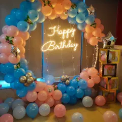 A classic circular balloon arch in a playful pink, blue, and peach color scheme. The "Happy Birthday" neon sign in the center adds a warm glow, making it a great backdrop for cake cutting and photos.