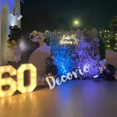 A grand 60th birthday celebration with a unique backdrop combining natural textures and modern lighting. The setup features large marquee numbers and a playful neon sign that reads 'Looks 40, Behaves 20', adding a fun, personal touch to the event.