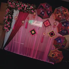 A full view of a Mehendi stage setup. The backdrop features a pink floral arch, a 'Mehendi' neon sign, and a wall decorated with colourful umbrellas and traditional hangings, all set on an artificial grass floor.