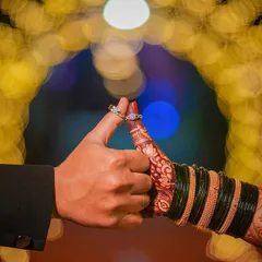 A beautiful and symbolic shot focusing on the couple's hands and rings. The bride's hand, adorned with mehndi and traditional green bangles, is gently held by the groom, showcasing their engagement rings.