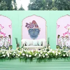 This Mehendi stage is designed around a Pichwai theme, featuring a custom backdrop with a traditional painting of Shrinathji. The seating area is framed with fresh white flowers and set against a pastel green structure.