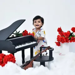 A charming studio session featuring a baby boy with a miniature grand piano. The set is decorated with red roses and fluffy clouds for a dramatic and adorable look.