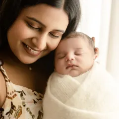 A close-up portrait of a new mother smiling as she holds her sleeping newborn. The soft lighting highlights the peaceful bond between them.