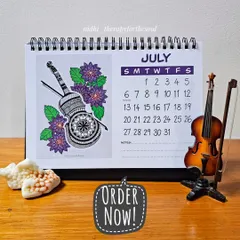 The July page of the calendar, showcasing a detailed violin illustration nestled among purple dahlias. The calendar is spiral-bound and sits perfectly on any desk.