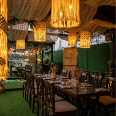 This rustic dining setup features a long wooden table under a canopy of draped lace and hanging macrame lanterns. The warm lighting and natural textures create an inviting and stylish setting for an alfresco dinner.